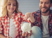 Pretty girl is putting a coin into a piggy bank that her handsome father is holding. Both are smiling while sitting on sofa at home
