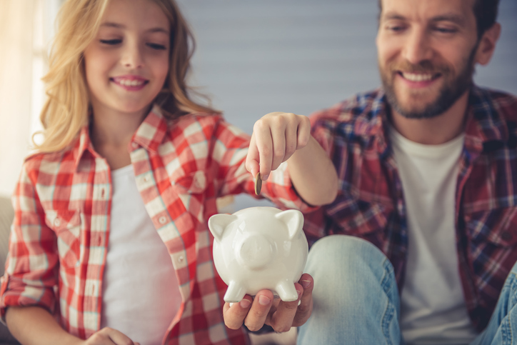 Pretty girl is putting a coin into a piggy bank that her handsome father is holding. Both are smiling while sitting on sofa at home